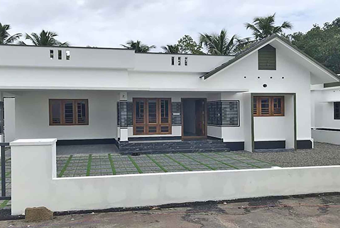 Front view of a contemporary 1400 sq. ft. white 3 BHK villa in a gated complex on Chembakassery Road, featuring a gabled roof section, wooden window frames, and a paved driveway with green accents.