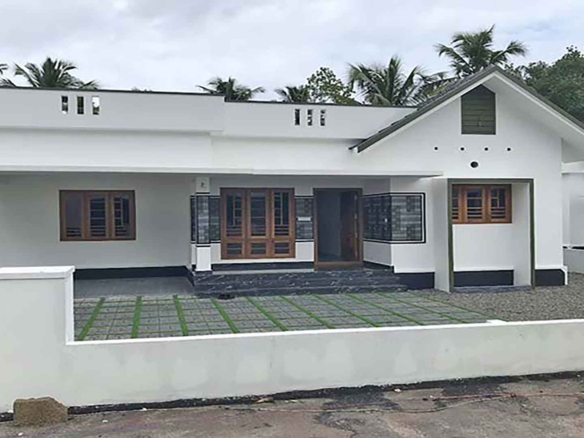Front view of a contemporary 1400 sq. ft. white 3 BHK villa in a gated complex on Chembakassery Road, featuring a gabled roof section, wooden window frames, and a paved driveway with green accents.