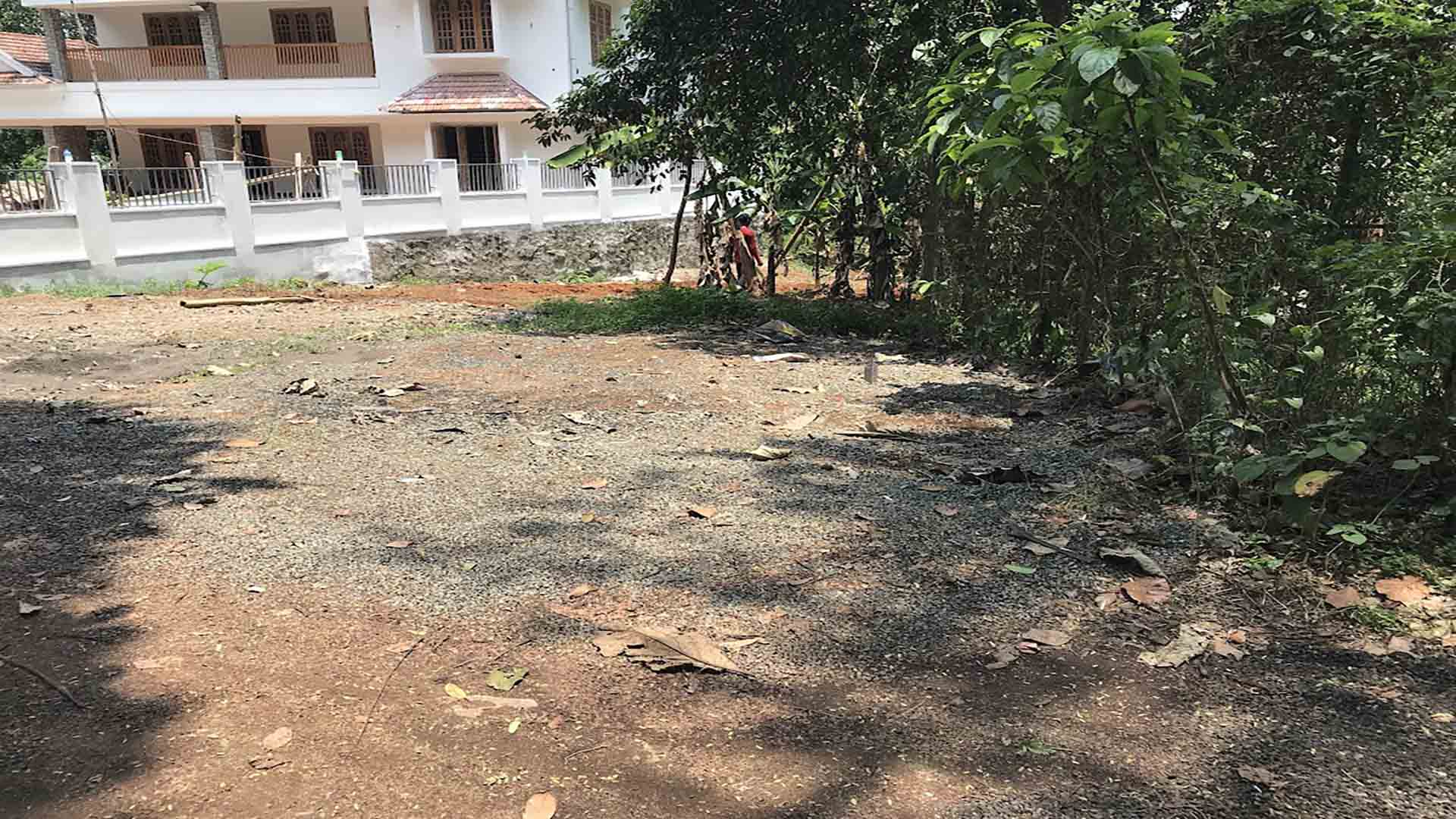 A cleared 21-cent residential land parcel for sale near TK Road, Thiruvalla, featuring level ground and a neighboring modern two-story house.