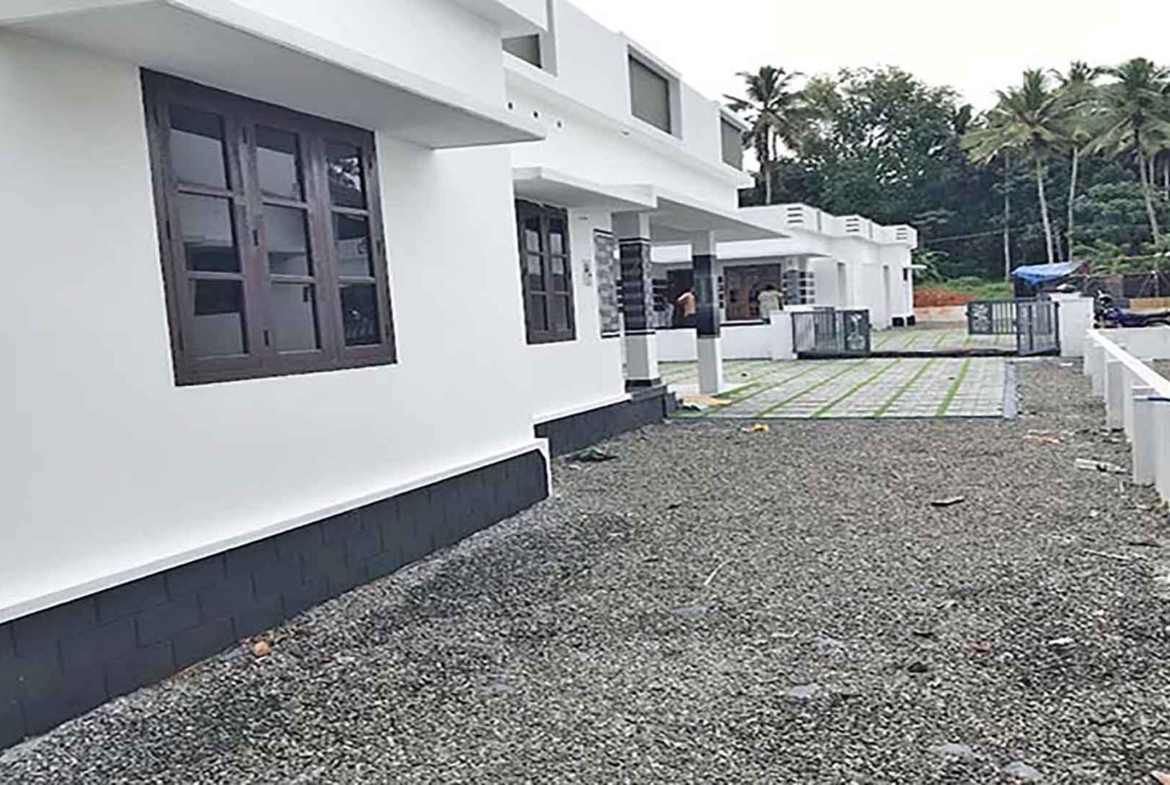 Perspective view of a modern white 3 BHK villa in a gated complex on Chembakassery Rd, featuring dark wood window frames, a grey stone foundation, and a gravel yard leading to adjacent villas.