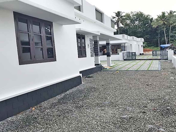 Perspective view of a modern white 3 BHK villa in a gated complex on Chembakassery Rd, featuring dark wood window frames, a grey stone foundation, and a gravel yard leading to adjacent villas.