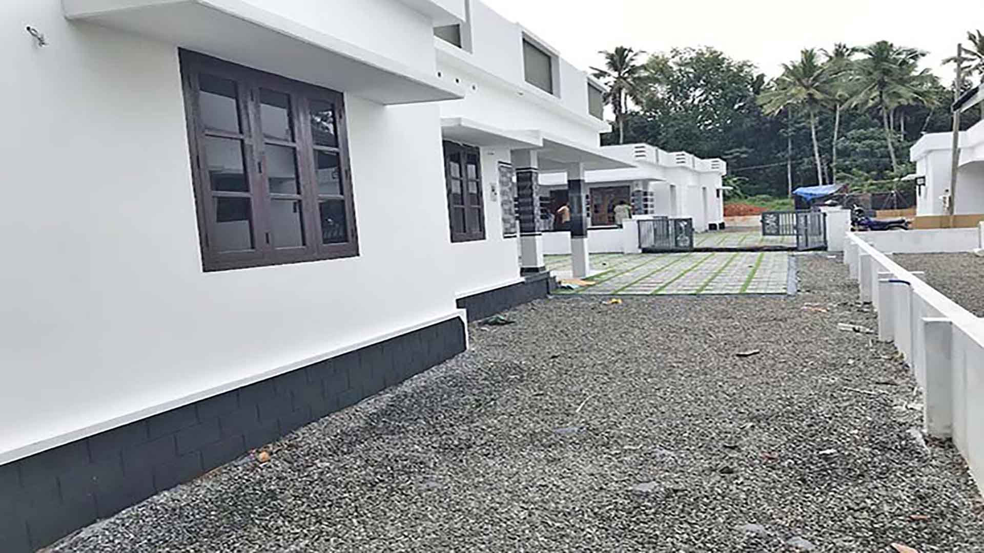 Perspective view of a modern white 3 BHK villa in a gated complex on Chembakassery Rd, featuring dark wood window frames, a grey stone foundation, and a gravel yard leading to adjacent villas.