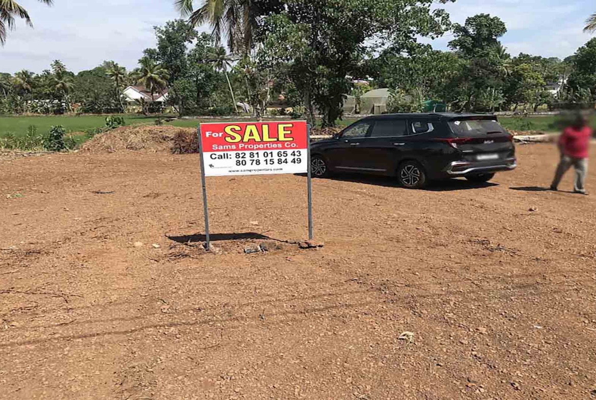 A cleared, level plot of land for sale in Pullad, Thiruvalla, featuring a red "For Sale" sign by Sams Properties Co. and a black SUV parked on the site against a backdrop of lush green coconut trees and paddy fields.