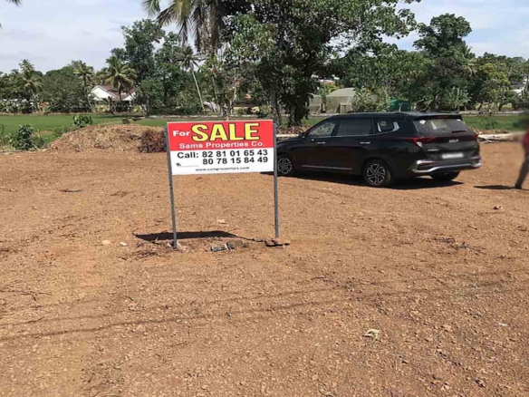 A cleared, level plot of land for sale in Pullad, Thiruvalla, featuring a red "For Sale" sign by Sams Properties Co. and a black SUV parked on the site against a backdrop of lush green coconut trees and paddy fields.