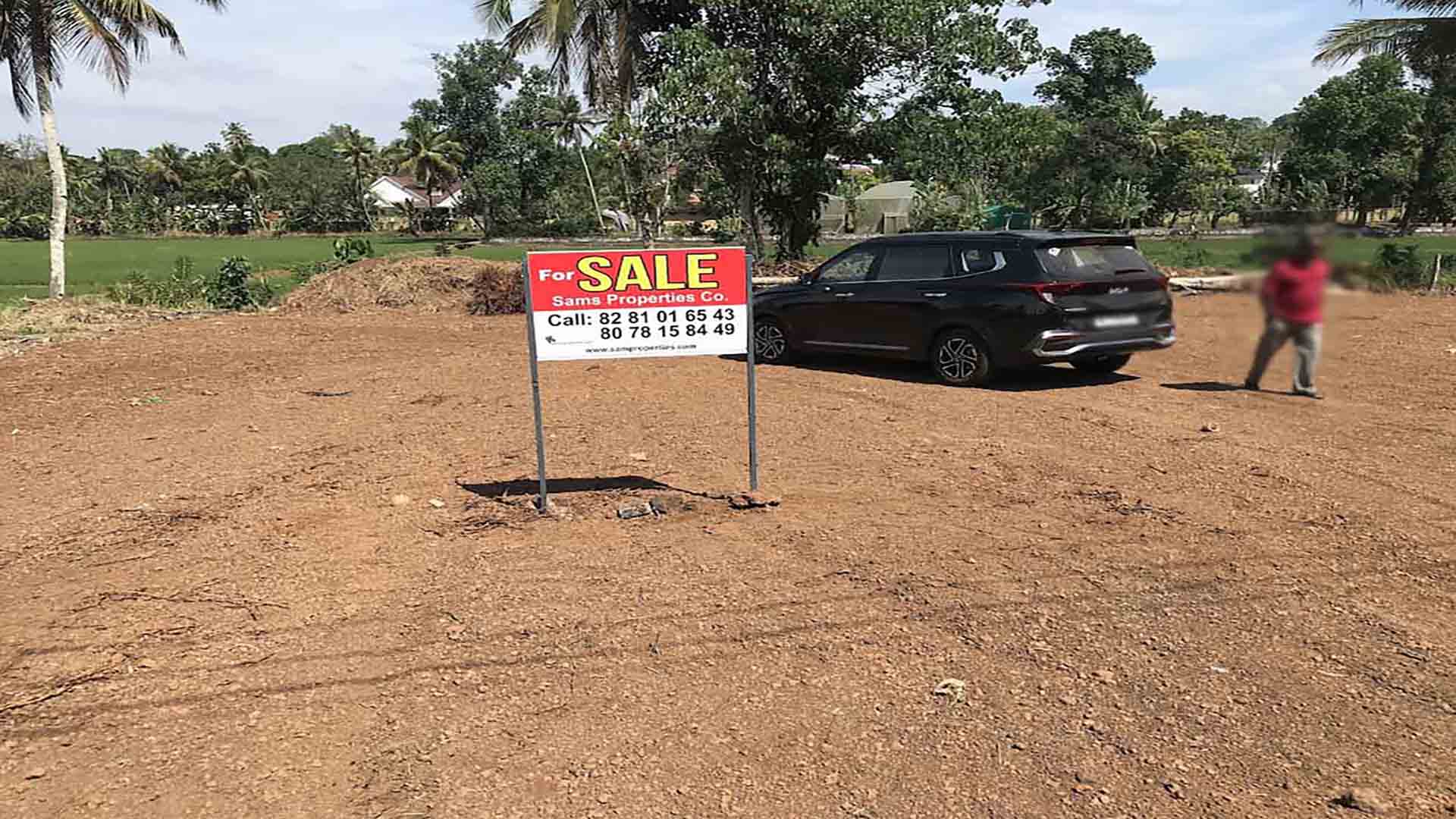 A cleared, level plot of land for sale in Pullad, Thiruvalla, featuring a red "For Sale" sign by Sams Properties Co. and a black SUV parked on the site against a backdrop of lush green coconut trees and paddy fields.