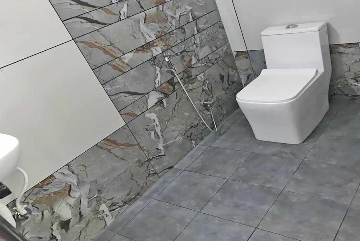 Modern bathroom interior featuring a white one-piece toilet against a decorative marble-patterned tile wall and grey floor tiles.