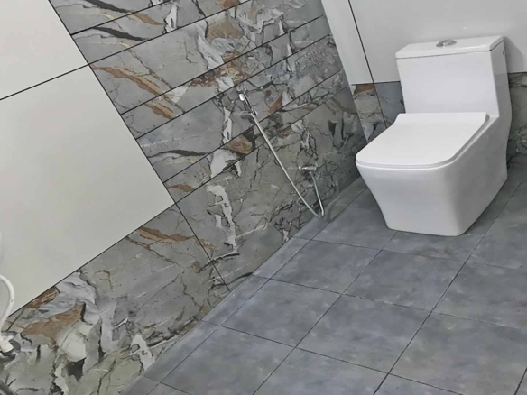 Modern bathroom interior featuring a white one-piece toilet against a decorative marble-patterned tile wall and grey floor tiles.