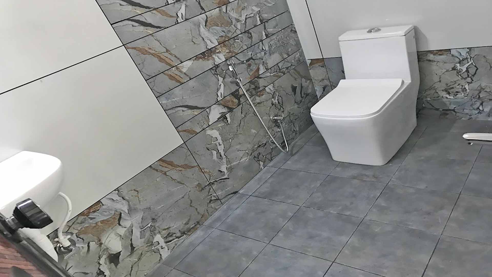 Modern bathroom interior featuring a white one-piece toilet against a decorative marble-patterned tile wall and grey floor tiles.