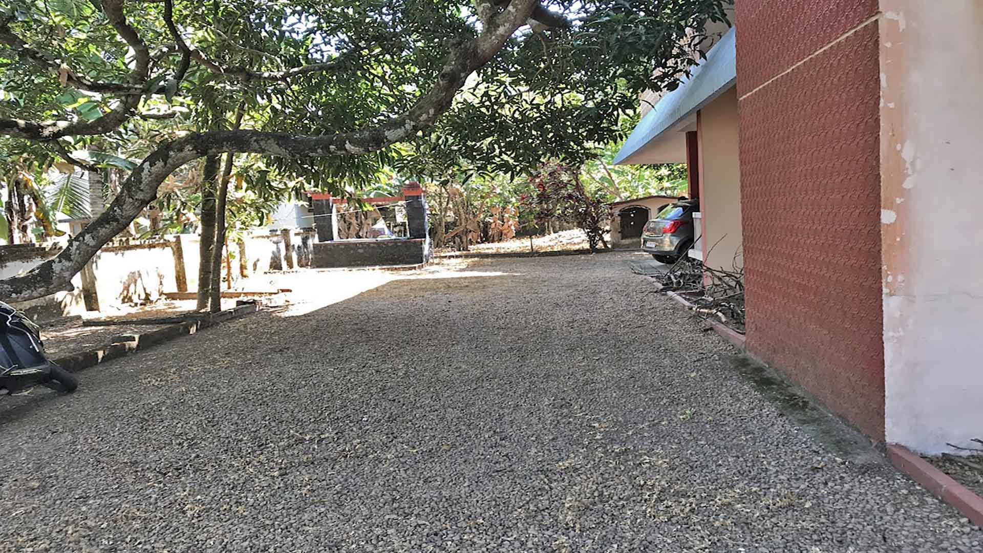 Wide gravel driveway and side view of a 4-BHK house on a 26-cent plot in Karthika Road, Chengannur, featuring mature mango trees, live water well and ample parking space.
