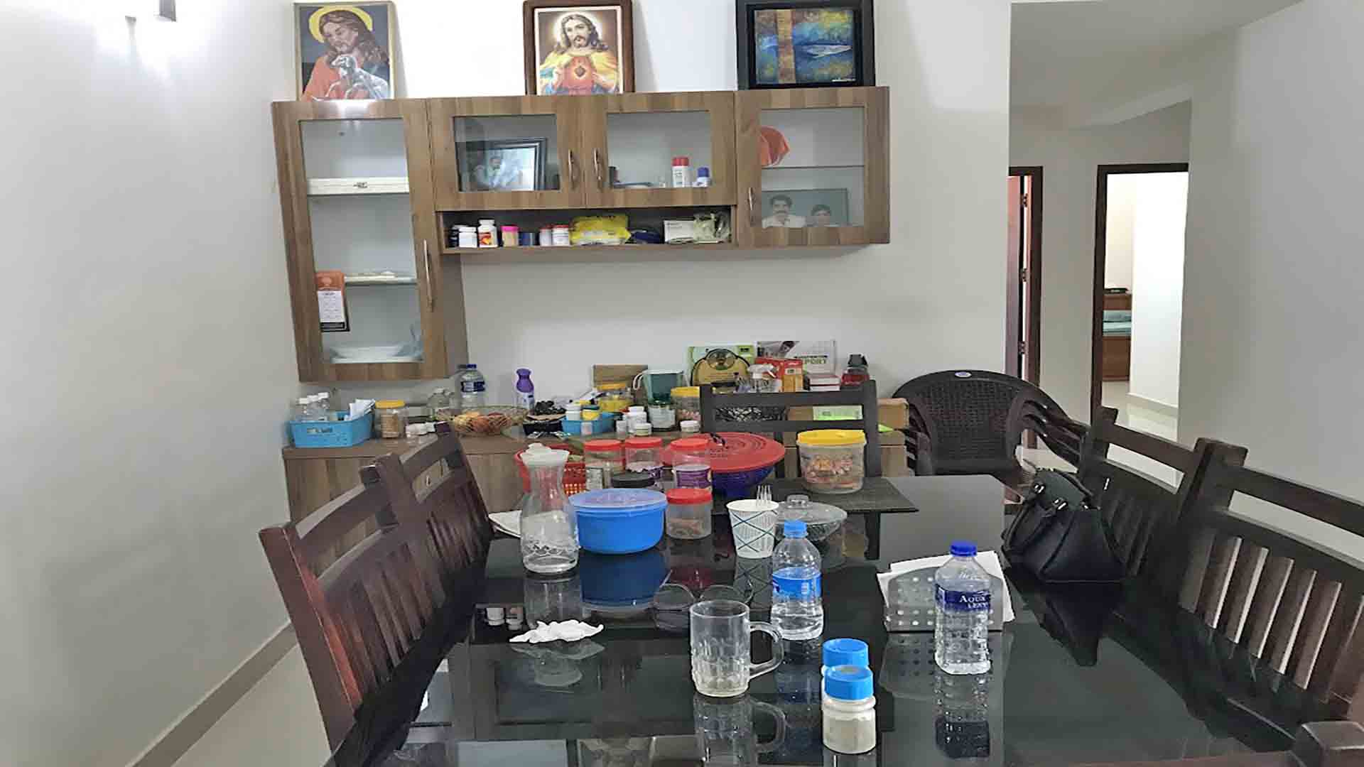 A modern dining room featuring a dark wood dining table with a glass top, matching wooden chairs, and a wall-mounted wooden crockery cabinet with glass doors.