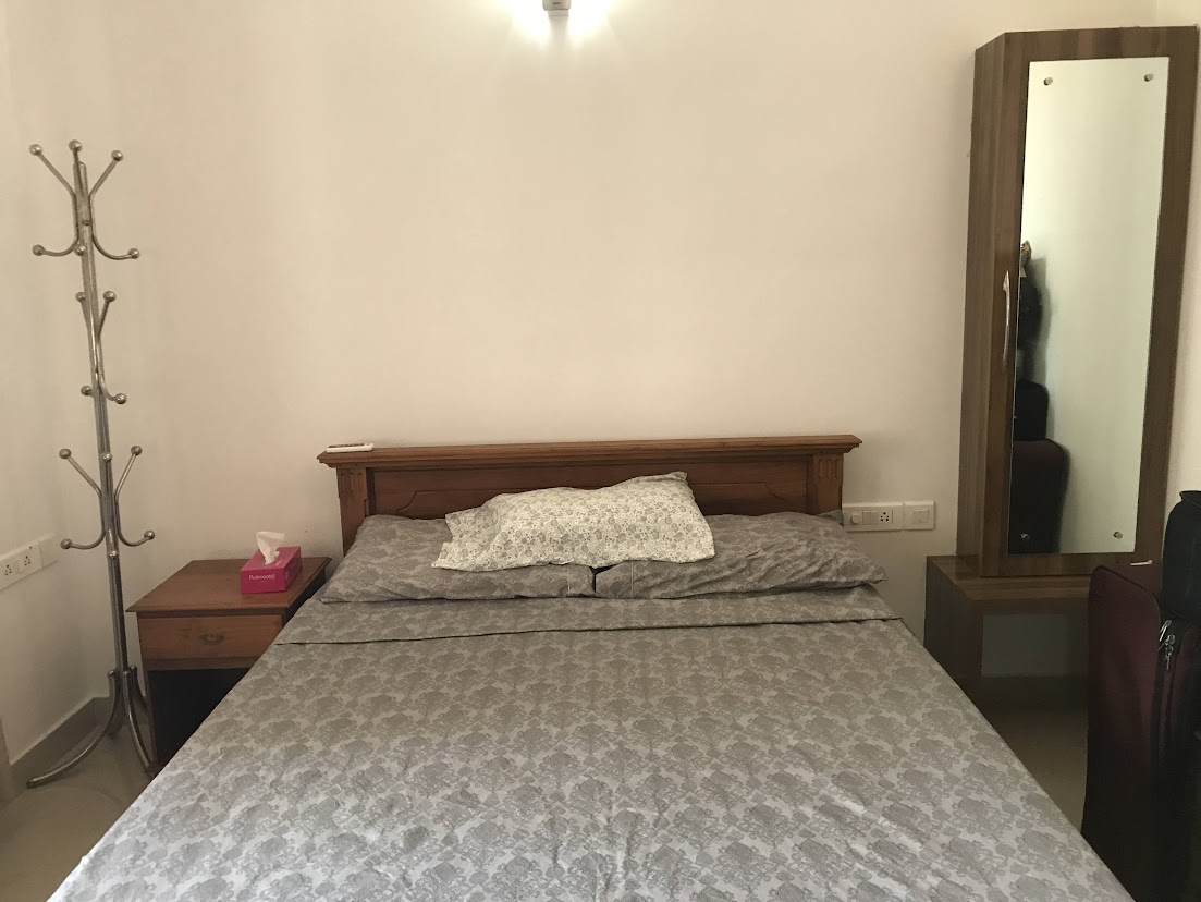 A fully furnished bedroom in a 3 BHK flat at The Edge by Skyline, Tiruvalla, featuring a wooden double bed, bedside table, standing coat rack, and a tall dressing mirror.