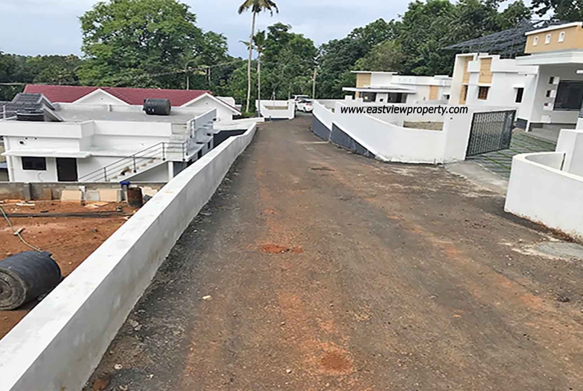 An elevated, wide-angle view of a private villa development in Kumbanad, Kerala, showing a sloped asphalt access road lined with white boundary walls, leading past modern white villas toward a lush forest and palm tree horizon under an overcast sky.