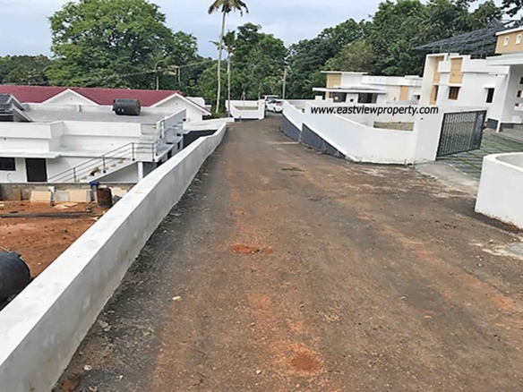 An elevated, wide-angle view of a private villa development in Kumbanad, Kerala, showing a sloped asphalt access road lined with white boundary walls, leading past modern white villas toward a lush forest and palm tree horizon under an overcast sky.