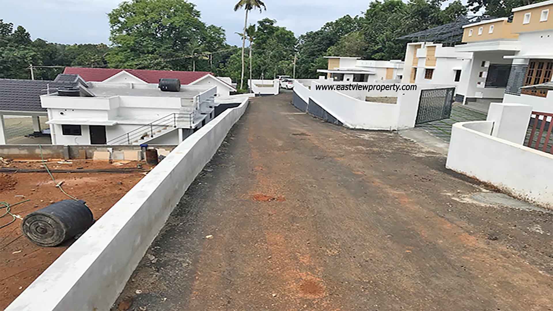 An elevated, wide-angle view of a private villa development in Kumbanad, Kerala, showing a sloped asphalt access road lined with white boundary walls, leading past modern white villas toward a lush forest and palm tree horizon under an overcast sky.