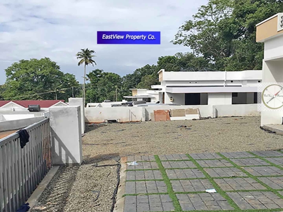 An elevated view of a modern gated villa complex in Thiruvalla, Kerala, showing multiple white villas with flat roofs, gray interlocking patio pavers with grass inserts, and a gravel driveway, all surrounded by lush tropical greenery and palm trees under a cloudy sky.