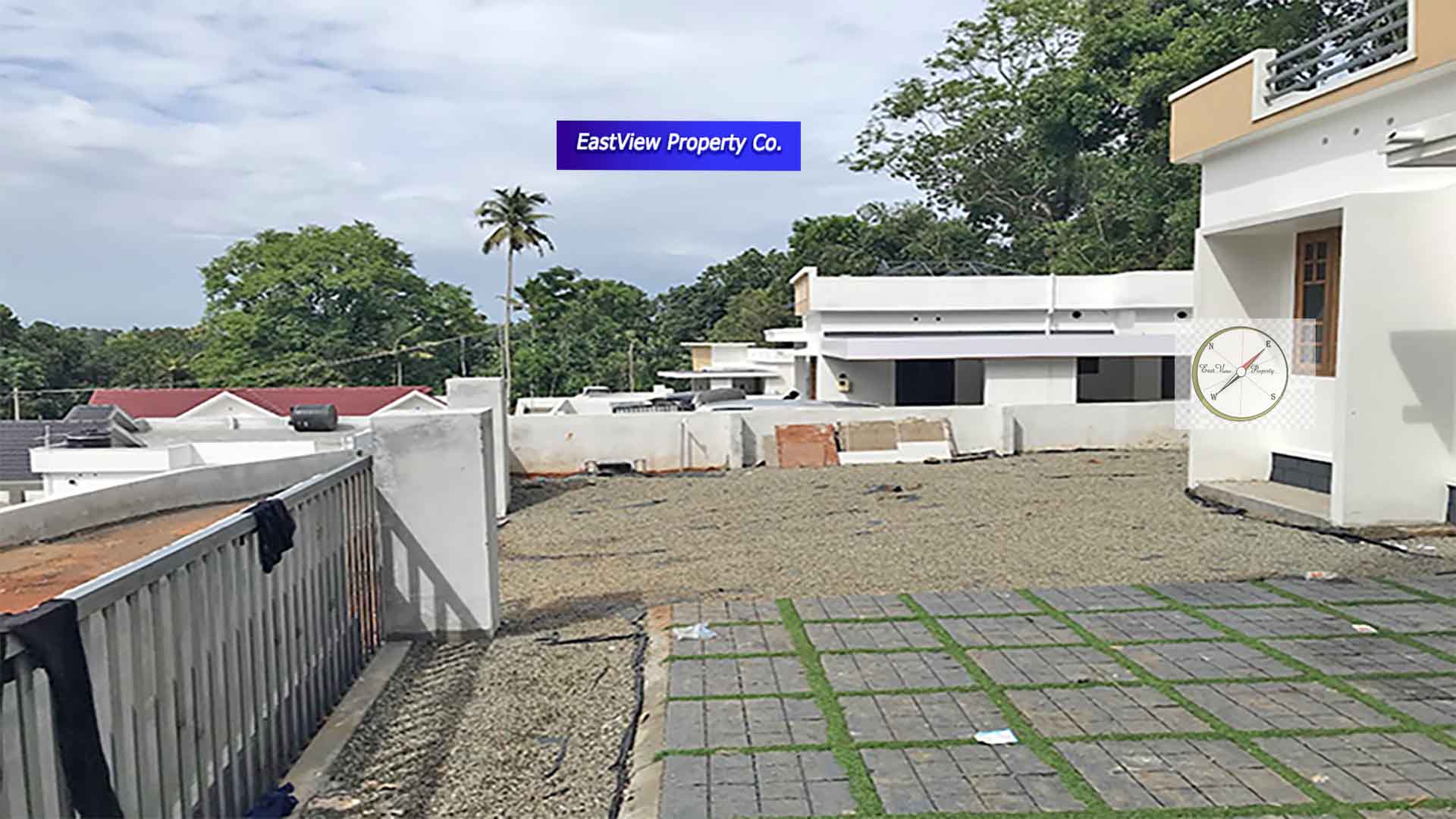 An elevated view of a modern gated villa complex in Thiruvalla, Kerala, showing multiple white villas with flat roofs, gray interlocking patio pavers with grass inserts, and a gravel driveway, all surrounded by lush tropical greenery and palm trees under a cloudy sky.