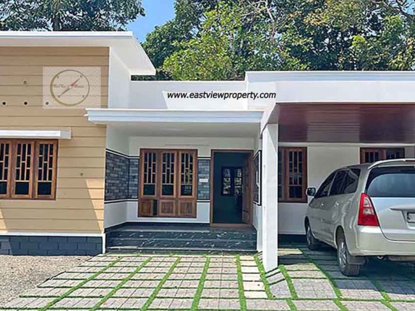 Exterior view of a modern single-story new 3BHK home in Thiruvalla, featuring a tan and white facade, a covered carport with a silver van, and a paved front yard with grass inserts.