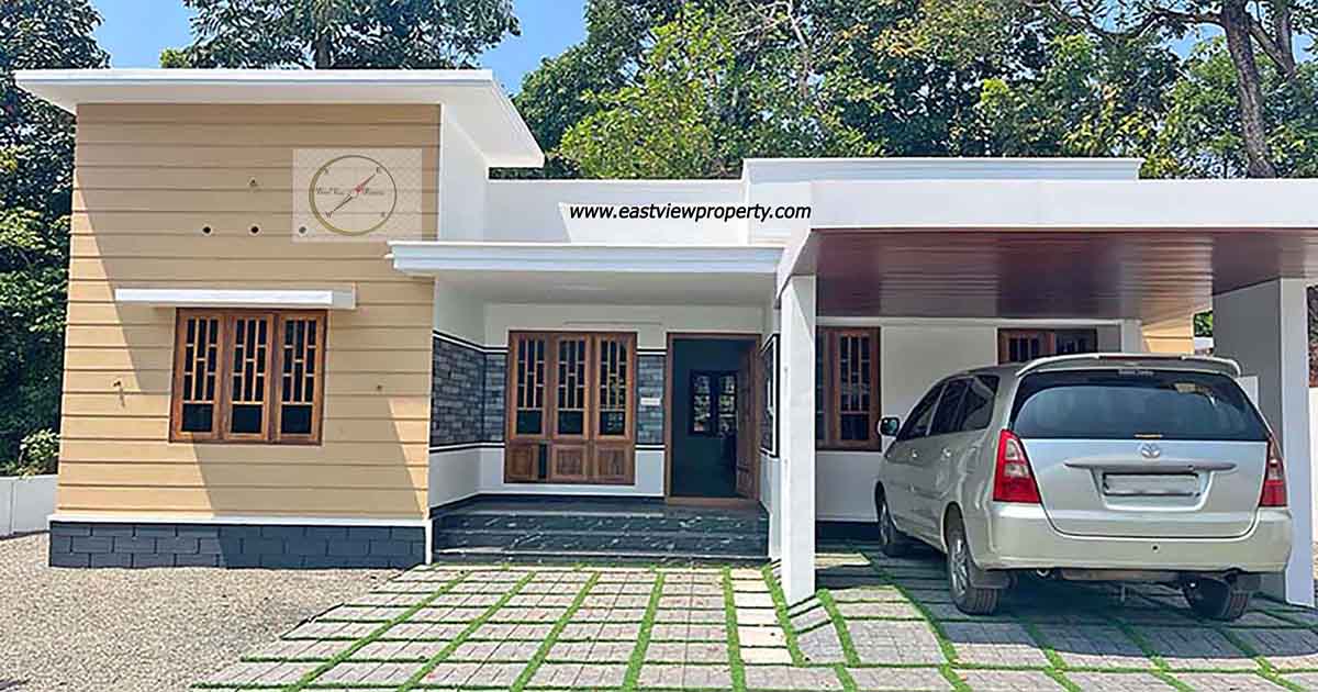 Exterior view of a modern single-story new 3BHK home in Thiruvalla, featuring a tan and white facade, a covered carport with a silver van, and a paved front yard with grass inserts.