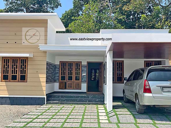 Exterior view of a modern single-story new 3BHK home in Thiruvalla, featuring a tan and white facade, a covered carport with a silver van, and a paved front yard with grass inserts.
