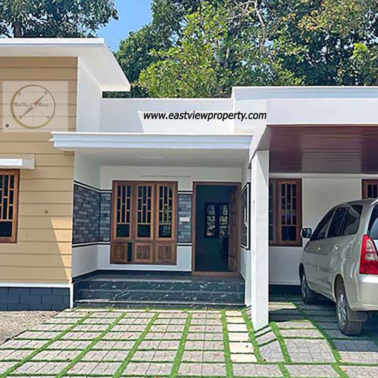 Exterior view of a modern single-story new 3BHK home in Thiruvalla, featuring a tan and white facade, a covered carport with a silver van, and a paved front yard with grass inserts.