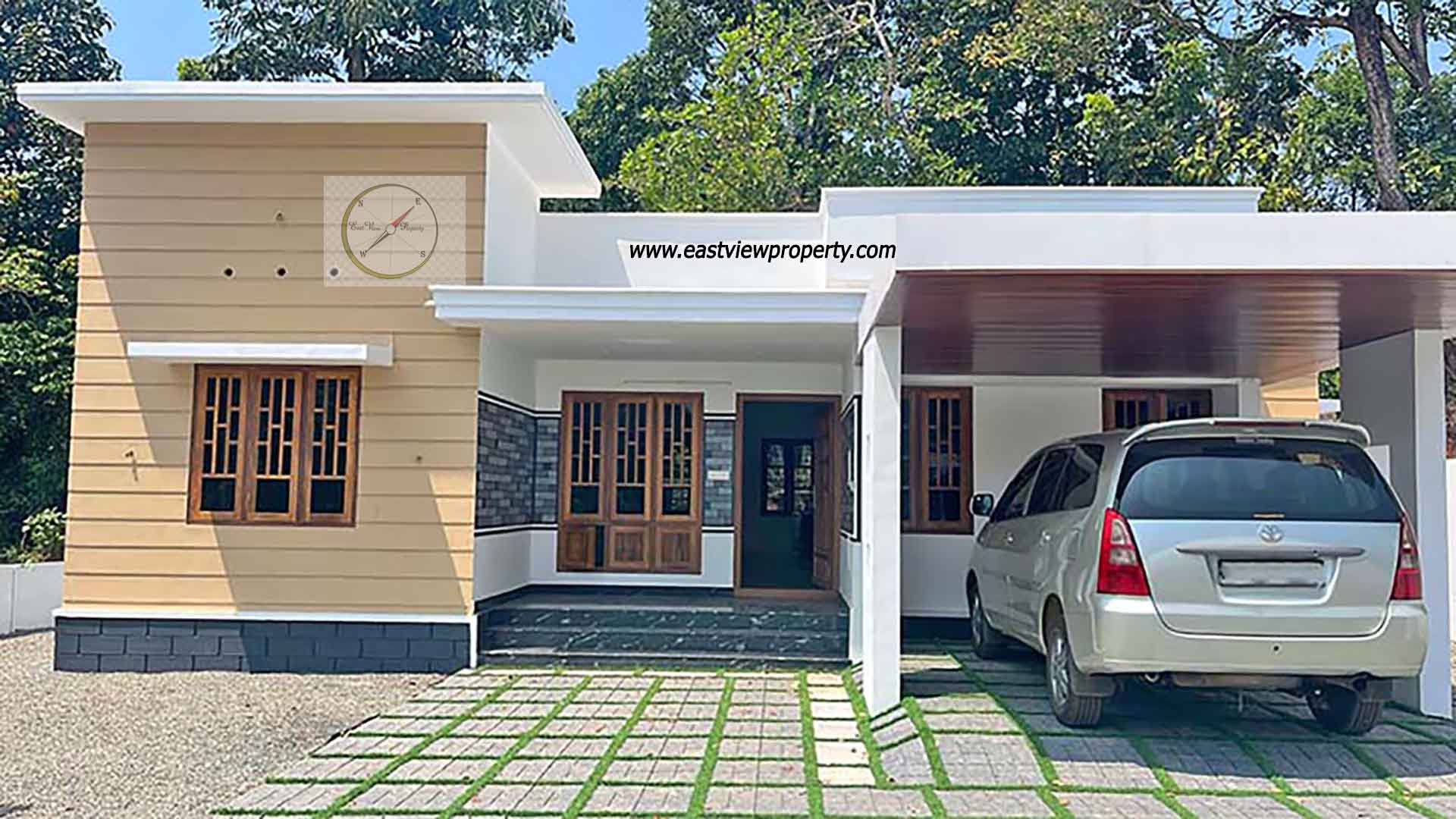 Exterior view of a modern single-story new 3BHK home in Thiruvalla, featuring a tan and white facade, a covered carport with a silver van, and a paved front yard with grass inserts.