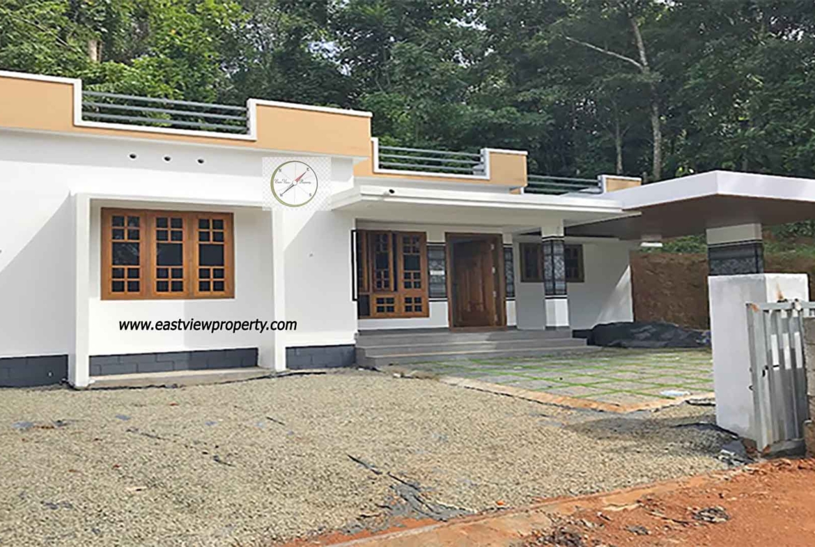 A modern single-storey white villa with a flat roof and mustard-yellow accents, featuring a spacious porch, a cantilevered carport, and wooden-framed windows, situated against a backdrop of dense green trees in Thiruvalla, Kerala.