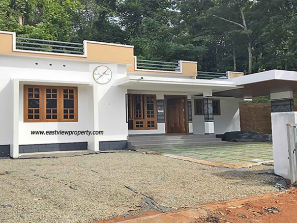A modern single-storey white villa with a flat roof and mustard-yellow accents, featuring a spacious porch, a cantilevered carport, and wooden-framed windows, situated against a backdrop of dense green trees in Thiruvalla, Kerala.