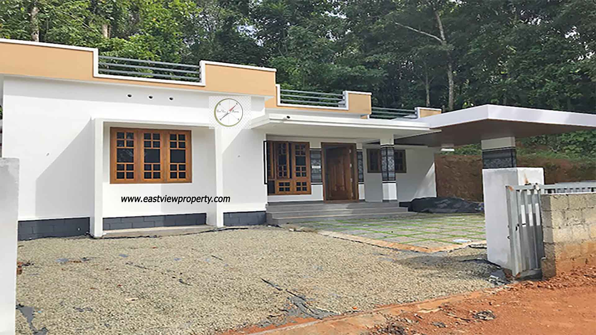 A modern single-storey white villa with a flat roof and mustard-yellow accents, featuring a spacious porch, a cantilevered carport, and wooden-framed windows, situated against a backdrop of dense green trees in Thiruvalla, Kerala.