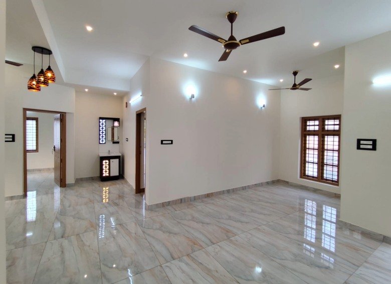 Large front living hall in a 3 BHK villa in Thiruvalla, featuring premium glazed vitrified flooring, false ceiling spot lighting, and dual ceiling fans.