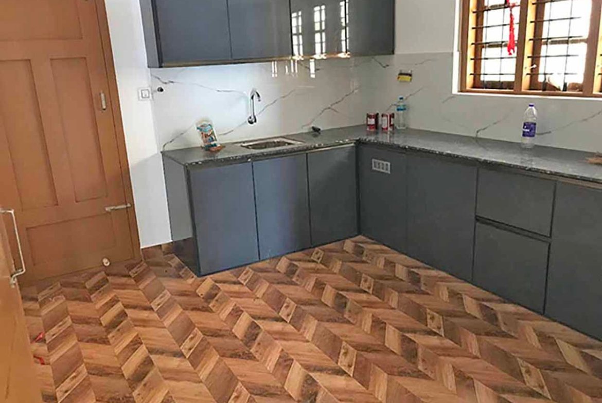 Modern L-shaped grey modular kitchen with dark granite countertop, stainless sink, and wooden herringbone floor tiles.