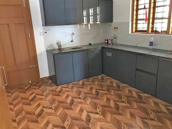 Modern L-shaped grey modular kitchen with dark granite countertop, stainless sink, and wooden herringbone floor tiles.